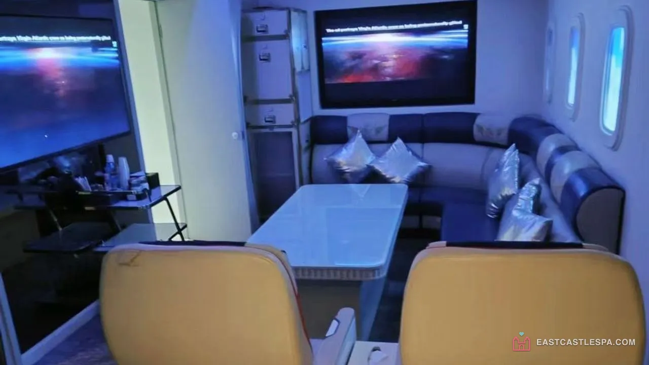 The aircraft cabin theme room at East Castle Spa, Macau sauna: two cream airline seats, a white table, a navy-and-white leather L-sofa with silver cushions, two screens and oval windows lit blue
